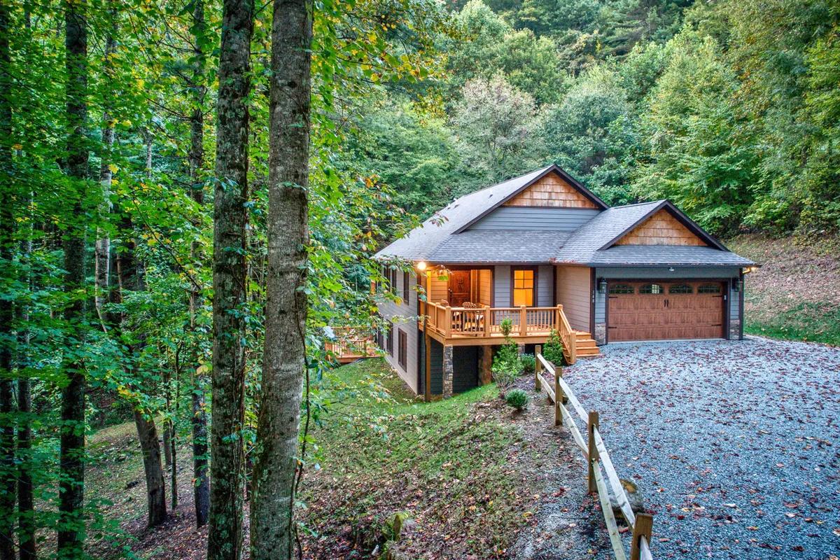 The image showcases the charming exterior of a newly built mountain cabin nestled among tall, peaceful trees. The inviting front porch, featuring cozy seating and an elegant wooden garage door, sets the scene for relaxing evenings spent watching sunsets or sipping coffee in the fresh mountain air.