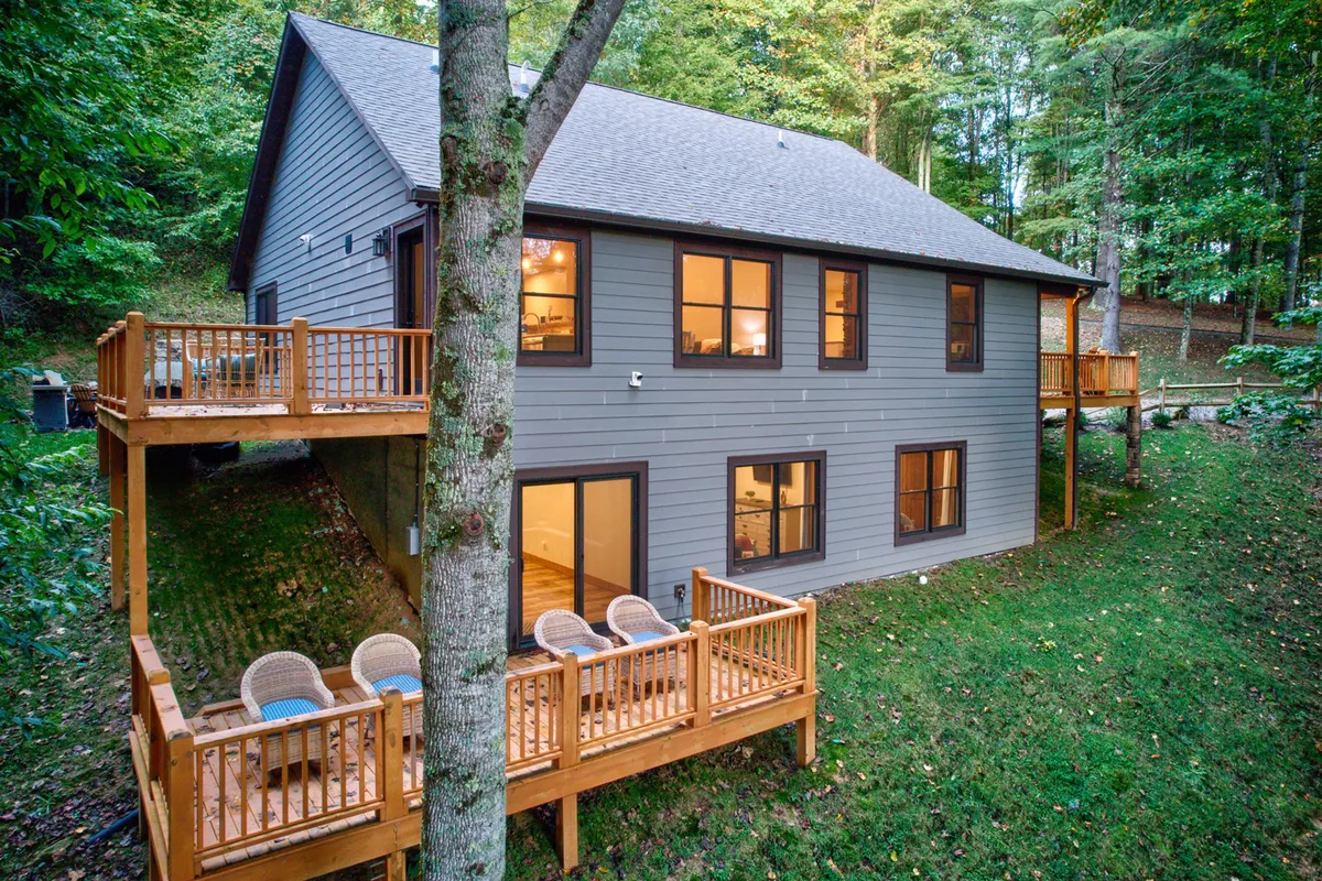 A wide shot of a wooden deck adjacent to large windows, with the surrounding trees and a grassy area visible. The deck is arranged for gatherings and relaxation, offering both sunlit and shaded spots for enjoying the mountain breeze. The tranquil scene invites guests to linger outdoors for coffee or cocktails while immersed in nature.