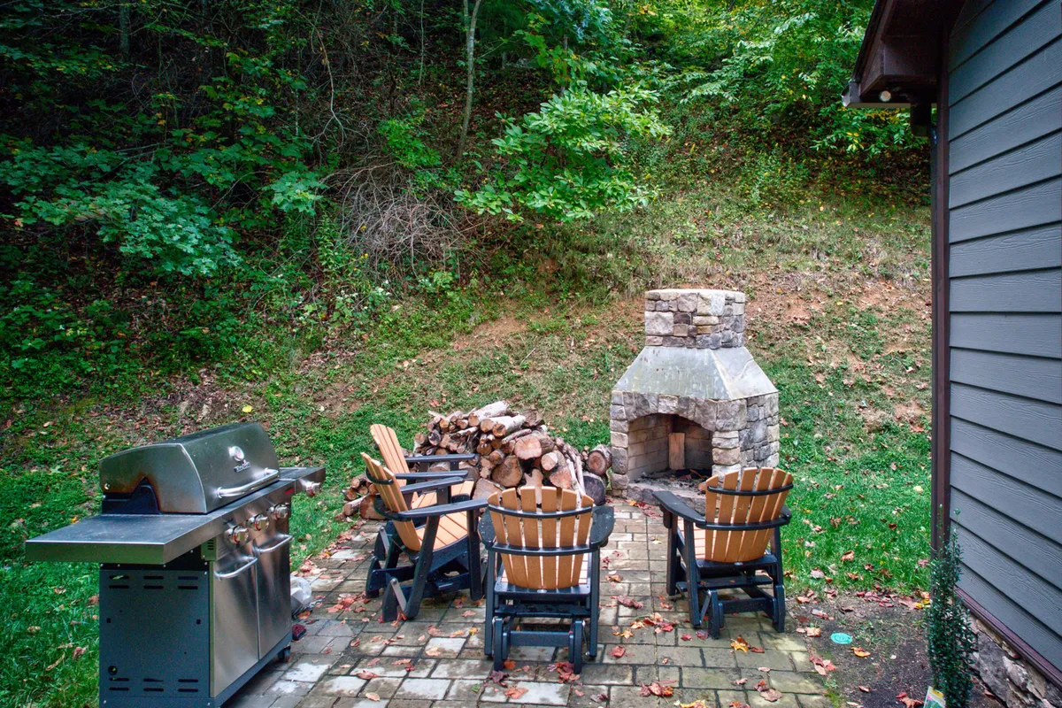 A close-up view of a paved patio area with a propane grill, stacked firewood, adirondack chairs, and a stone fireplace. The setting is designed for both cooking and social gatherings, surrounded by natural beauty. The arrangement highlights the versatility of the outdoor space for both alfresco dining and cozy evenings by the fire.
