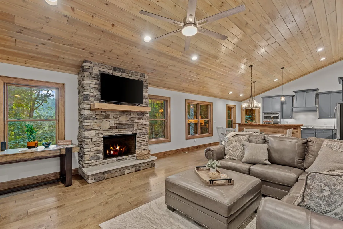 A spacious living room featuring a striking stone fireplace, large windows framing the outdoors, plush seating arranged for gatherings, and a dramatic wooden vaulted ceiling with tongue-and-groove accents. The natural light and rustic elements create a warm, inviting atmosphere ideal for family and friends.