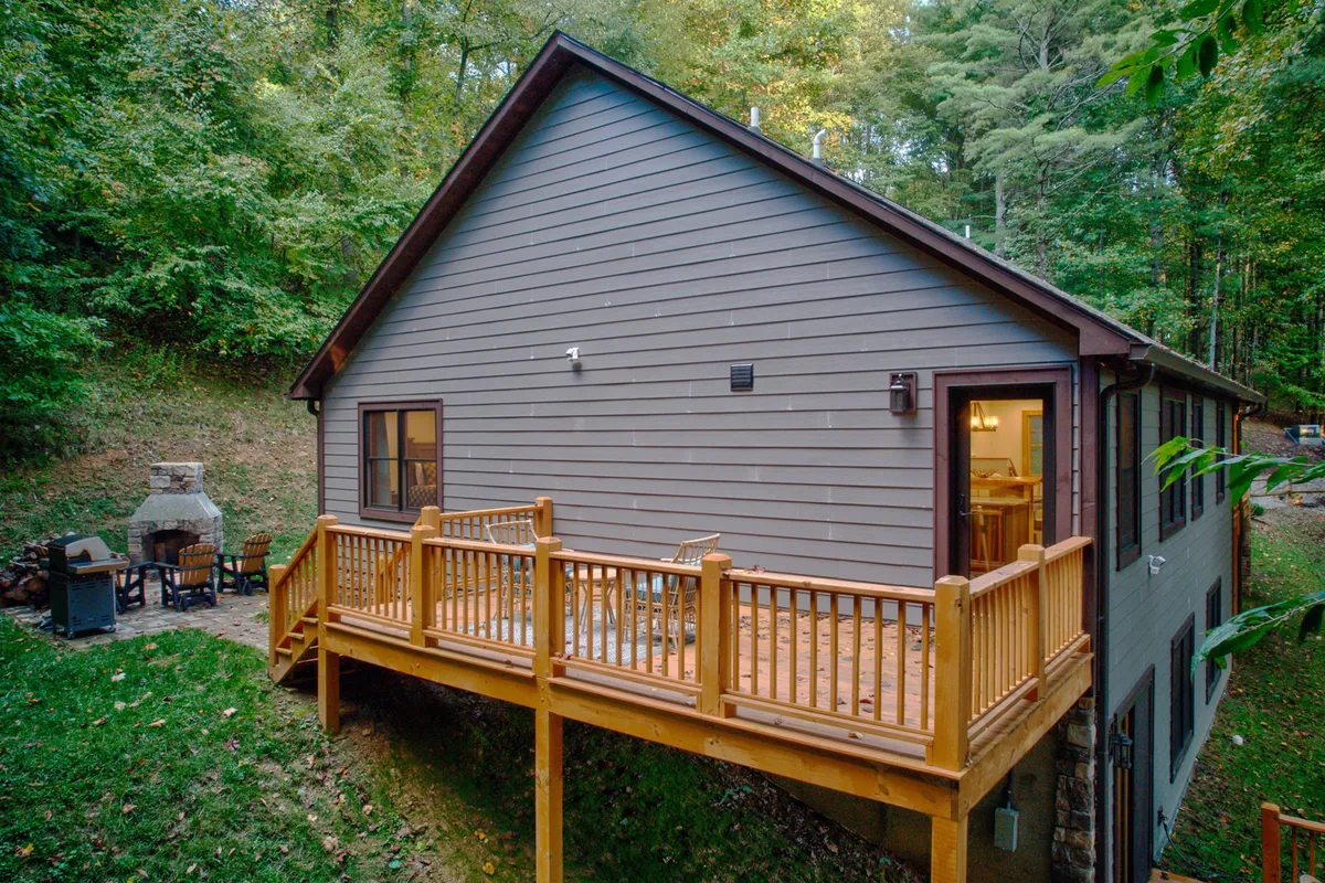 A spacious wooden deck set against the backdrop of surrounding trees, featuring a built-in outdoor fireplace, comfortable seating, and a dining area. The scene is set during golden hour, with the warm glow of sunset enhancing the inviting atmosphere. The deck is clearly designed for relaxation and socializing, with ample space for gatherings.