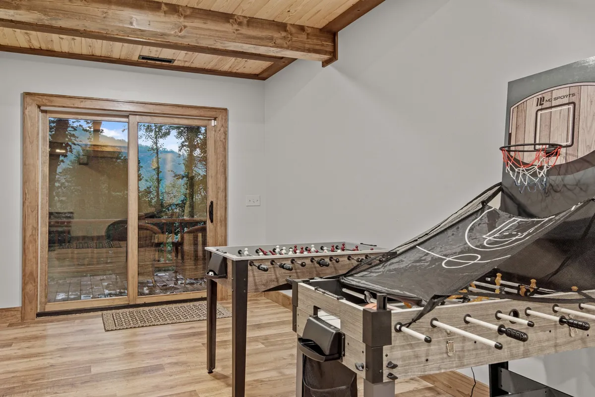 A vibrant game room featuring a foosball table, basketball arcade game, and a large sliding glass door that lets in natural light. The space is designed for interactive play and energetic family gatherings.