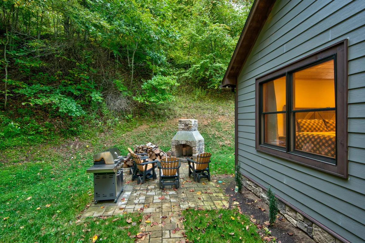 This image features a welcoming outdoor space with a stone fireplace, comfortable seating area, grill, and a window aglow with warm indoor light. The surrounding trees and relaxed setup evoke a sense of intimate connection with nature and cozy mountain evenings.
