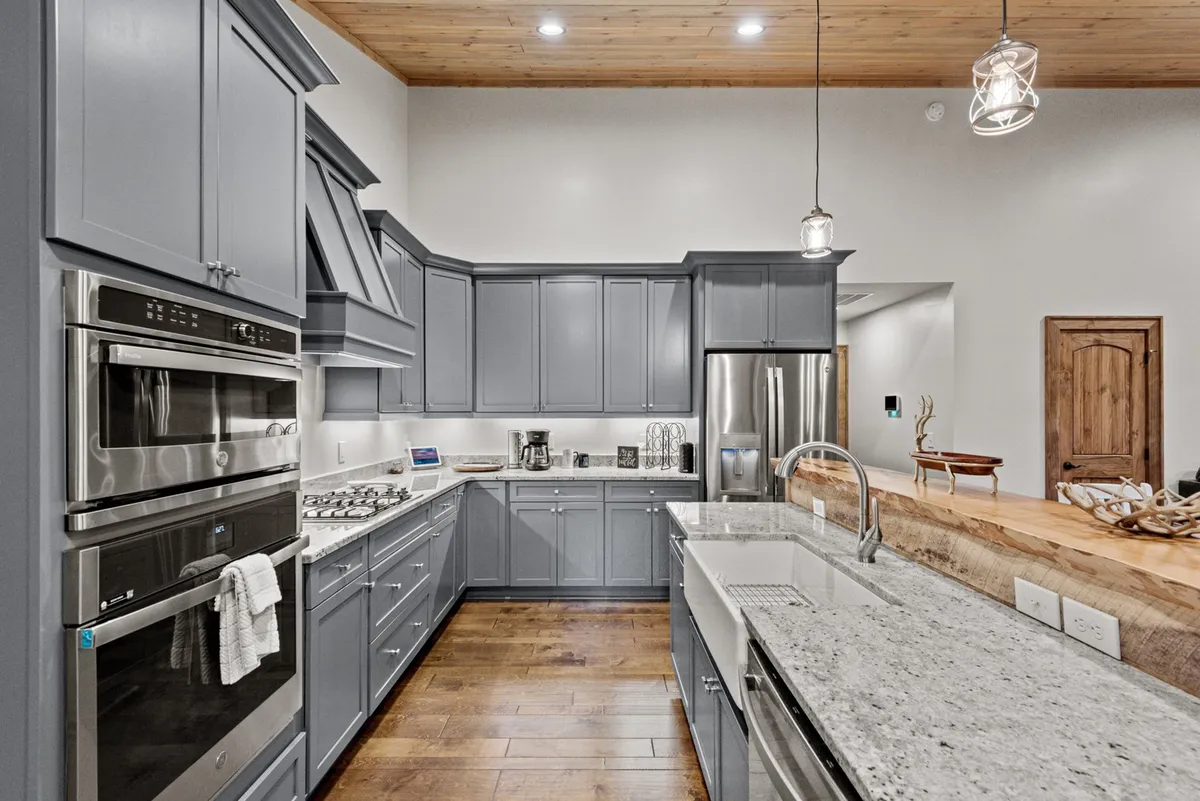 A modern kitchen with gray cabinetry, gleaming granite countertops, stainless steel appliances including a double oven, and pendant lighting. The wooden ceiling overhead ties the space to the rest of the open-concept design.