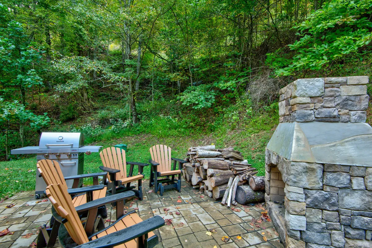 This image features a rustic outdoor stone fireplace with wooden chairs and a grill, surrounded by a stack of firewood. The setting invites groups to gather, cook, and enjoy stories or s’mores by the fire under the open sky.
